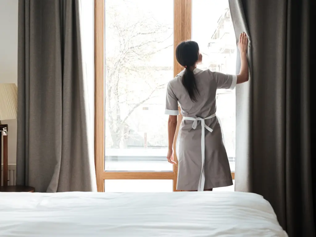 Housekeeping-Mitarbeiterin öffnet in einem hellen Hotelzimmer die Vorhänge, im Vordergrund das gemachte Bett